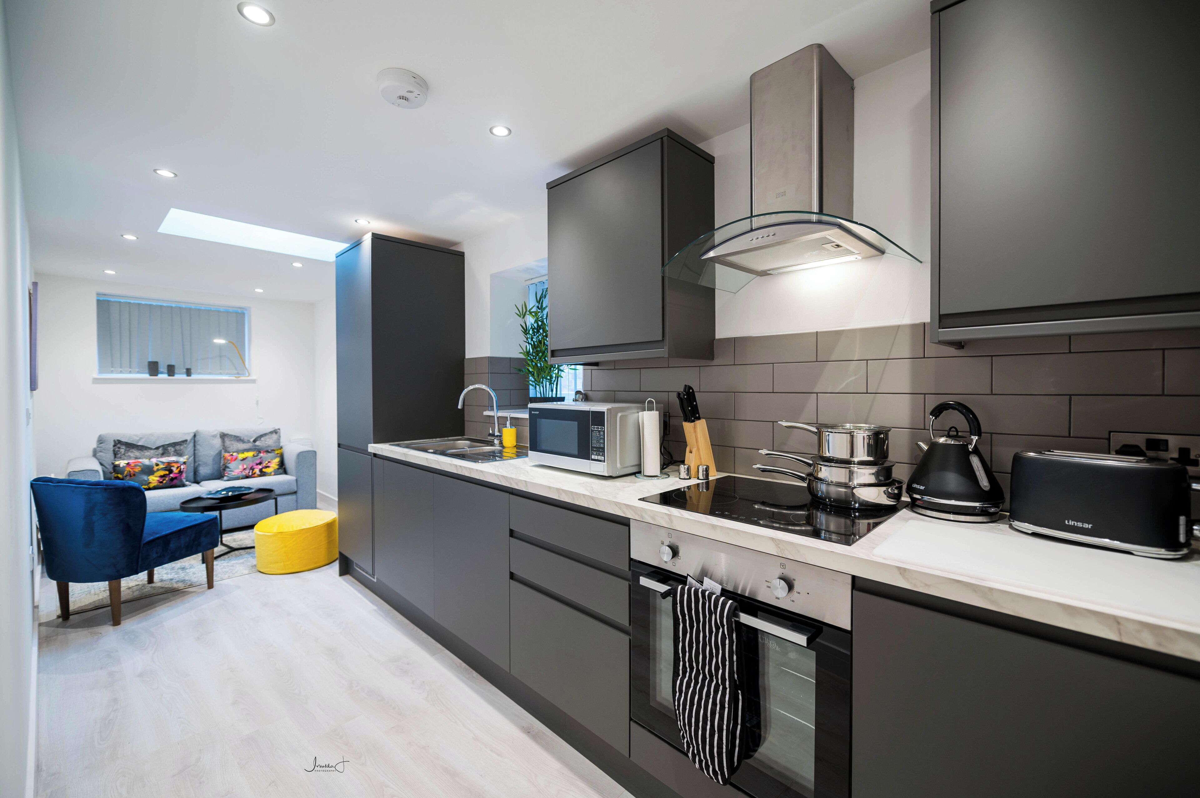 A modern kitchen with a built-in oven, microwave, and toaster. There is a dining area with a blue chair and a yellow ottoman.
