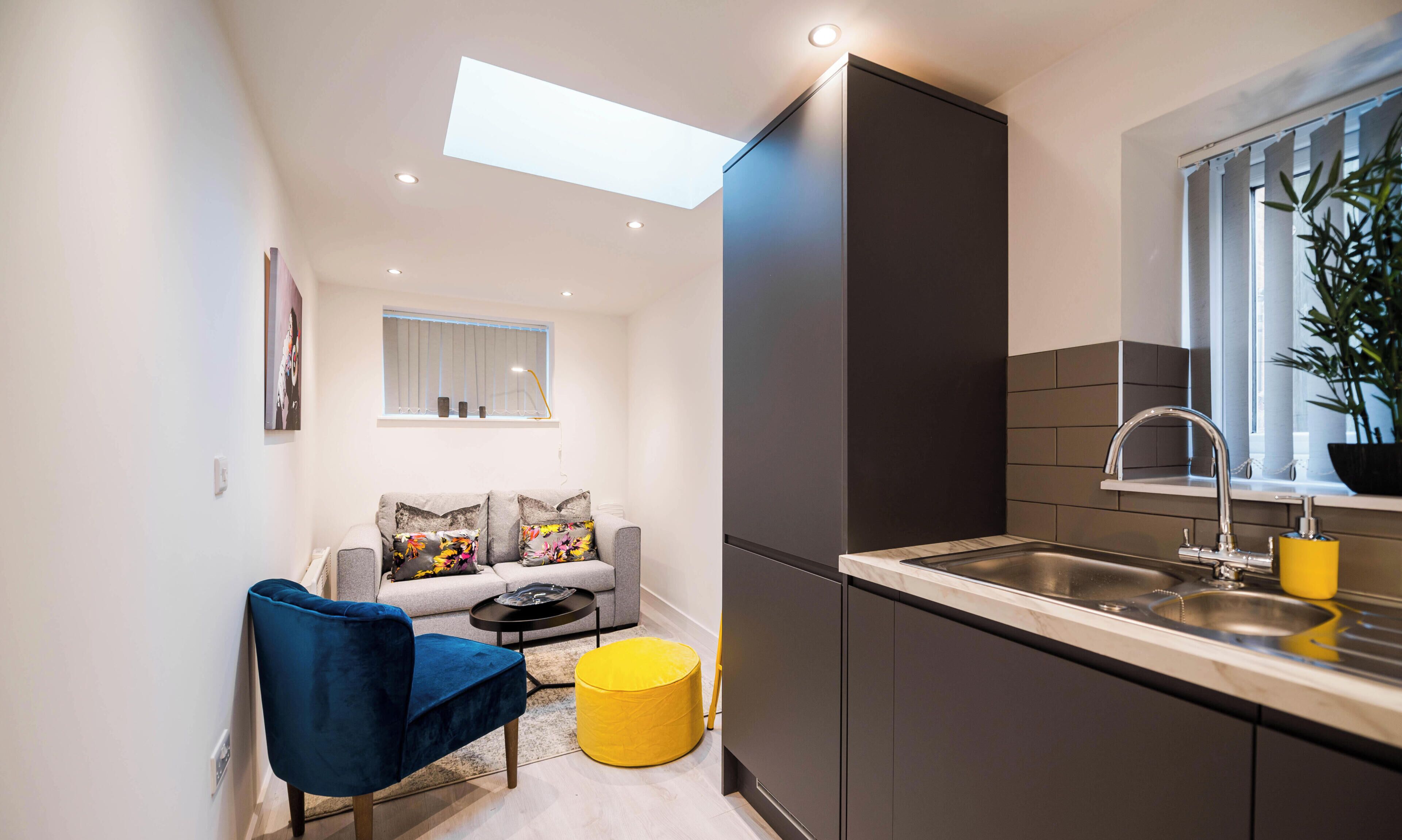 Een ruimtelijk ingerichte leefruimte met een kitchenette, een bank met bloemenkussens, een blauwe stoel, een geel voetenbankje en een raam met jaloezieën.