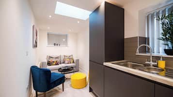 Een ruimtelijk ingerichte leefruimte met een kitchenette, een bank met bloemenkussens, een blauwe stoel, een geel voetenbankje en een raam met jaloezieën.