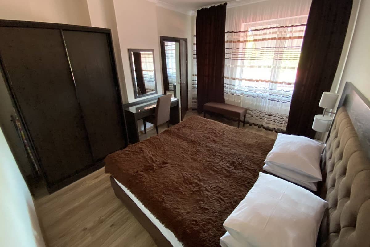 A bedroom with a large bed, a desk, a chair, a wardrobe, and a window with curtains.