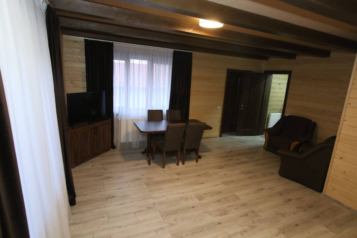 A room with a wooden floor, a dining table with chairs, a television on a wooden cabinet, and a dark-colored armchair.