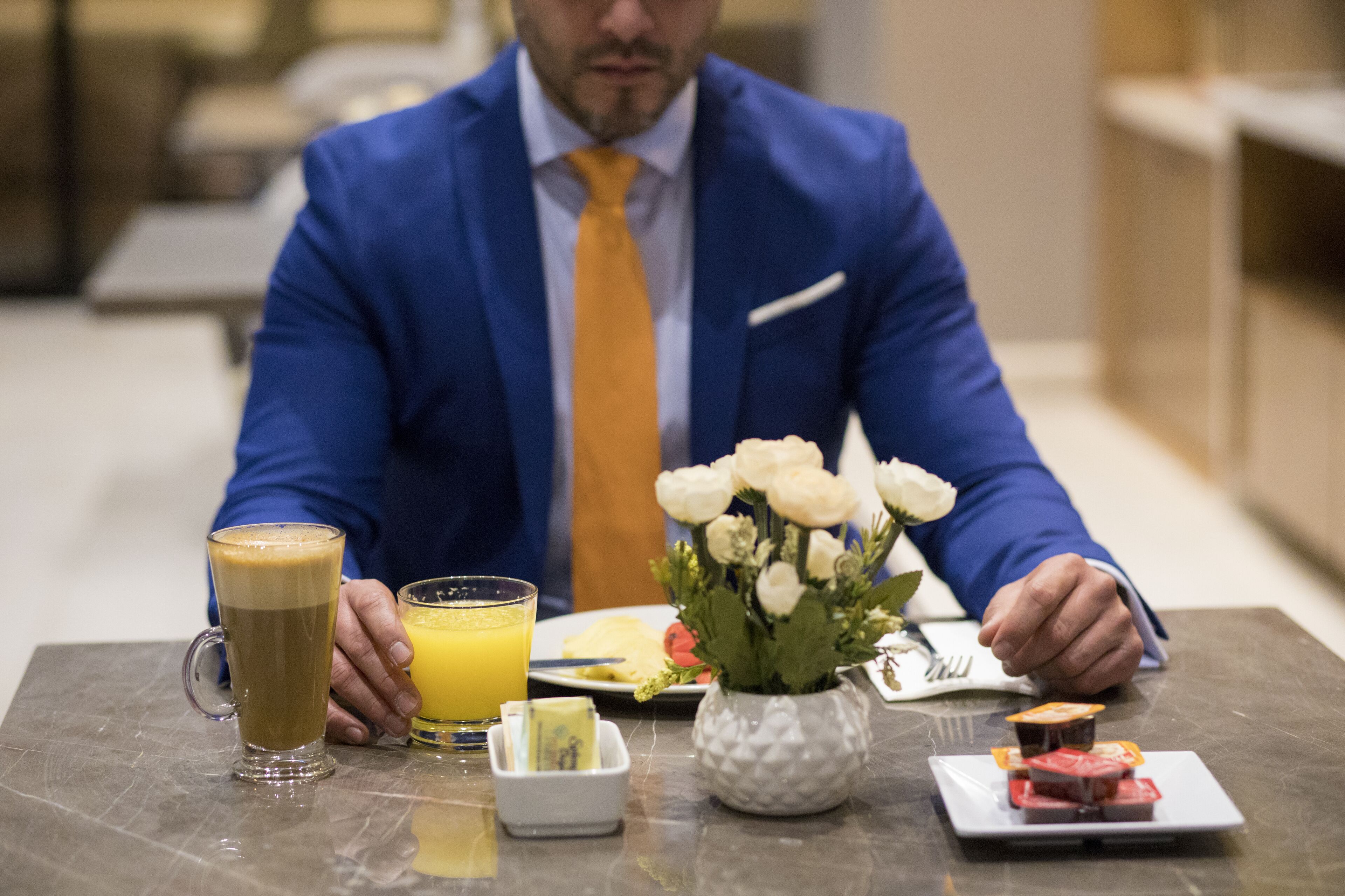 A man in a blue suit and yellow tie sitting at a table with a coffee mug, a glass of orange juice, a plate with food, and a vase with white flowers.