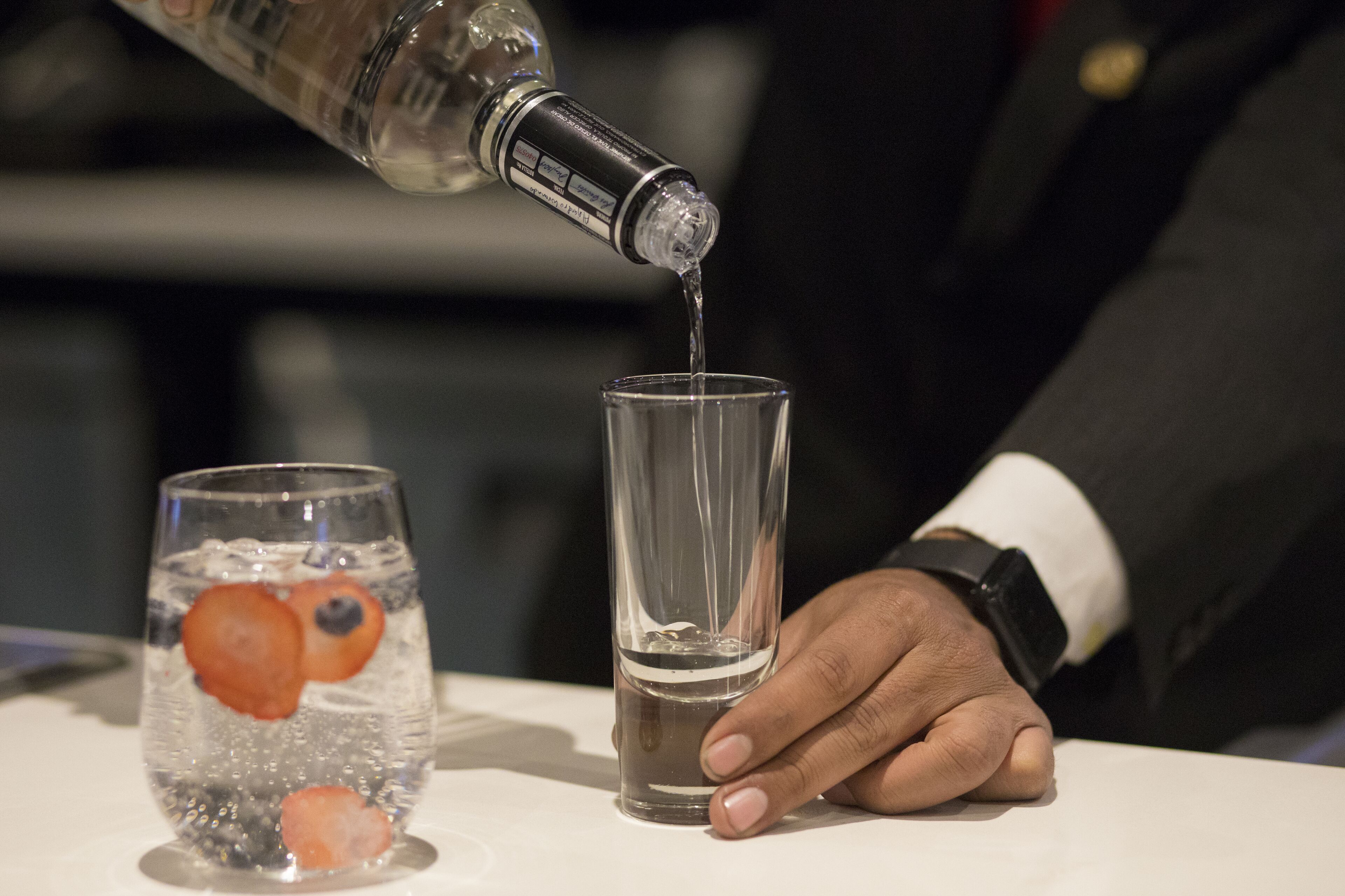 Una persona vertiendo un líquido transparente en un vaso, con una botella y un vaso con fruta y hielo sobre una mesa.
