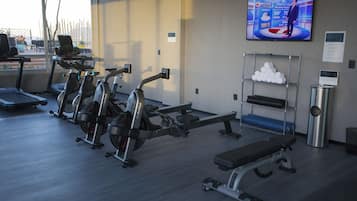 Academia com máquinas de cardio, bancos de peso e uma TV exibindo a previsão do tempo.