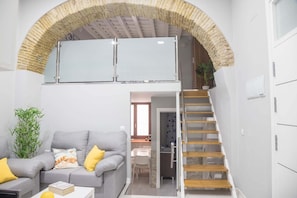 A compact living space with a grey sofa, a small table, a staircase, and a kitchen area.