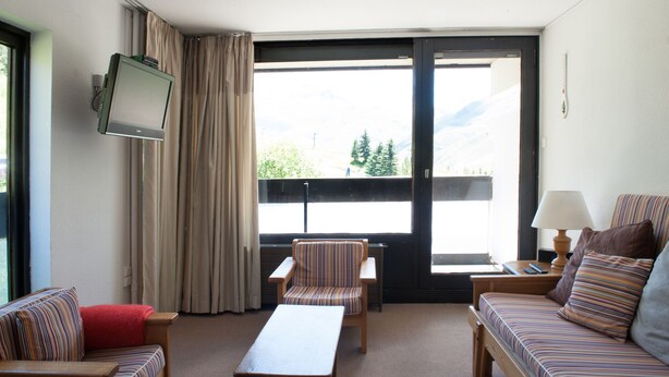 A hotel room with a sofa, a chair, a small table, a TV mounted on the wall, a window with a view of mountains, and a lamp on the side table. - Croisette / Dorons / 6 places (LES MENUIRES)