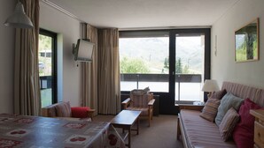 A hotel room with a bed, a sofa, a table, and a chair. There is a window with curtains and a view of the outdoors. - Croisette / Dorons / 6 places (LES MENUIRES)