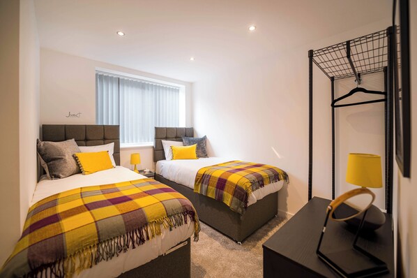 A hotel room with two beds, a window with blinds, a hanging clothes rack, and a bedside table with a lamp.