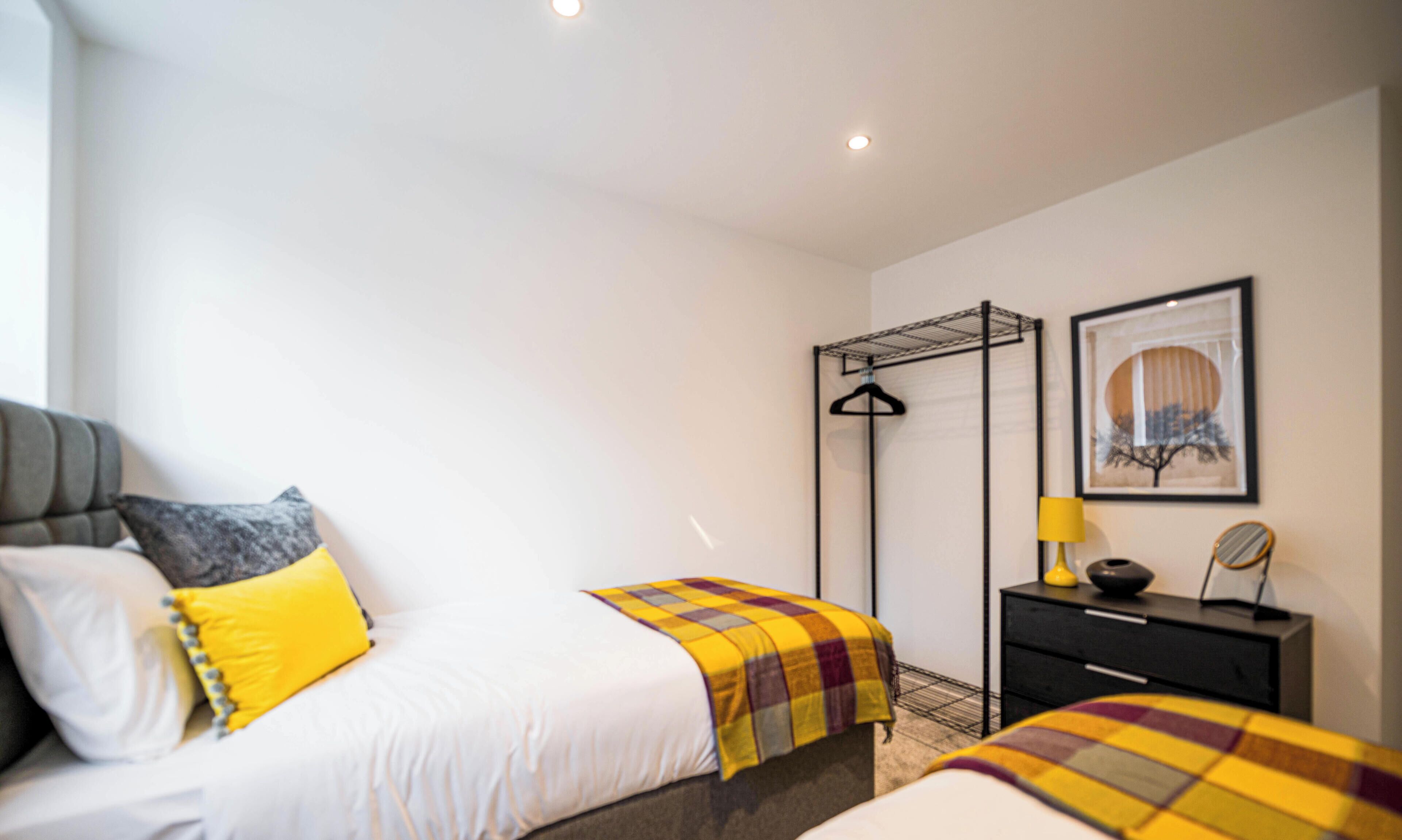A hotel room with a bed, a bedside table, a hanging clothes rack, and a framed artwork on the wall.