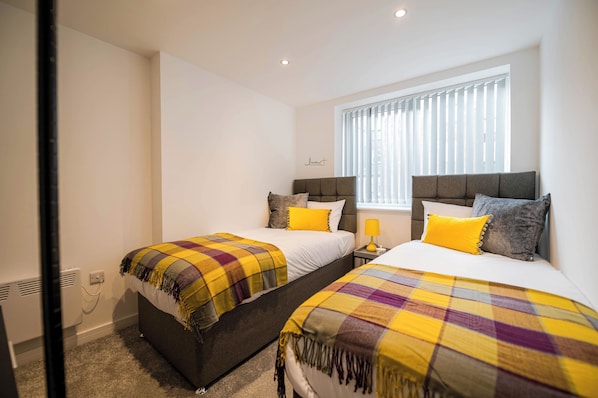 A bedroom with two beds, each with a headboard, a window with blinds, and a wall-mounted heater.