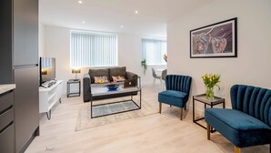 Een moderne woonkamer met een bank, een salontafel en een blauwe fauteuil. Aan de muur hangt een schilderij en op een bijzettafeltje staat een plant.