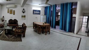 A spacious living room with a large sofa set, a dining table with chairs, a coffee table, and a patterned rug. There are curtains on the windows, and a ceiling fan is visible. - ADB Rooms Knights Inn (New Delhi)