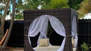 A wicker four-poster canopy bed with a beige mattress and pillows, set on a brick patio with a black fence and trees in the background. - Miami Skyline Luxury Suite With Private Patio and Parking . (Miami)