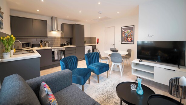A modern living room with a grey sofa, blue velvet chairs, a flat-screen TV, and a dining area with a white table and chairs. - Windmill House Apartment 4 (Lytham Saint Annes)