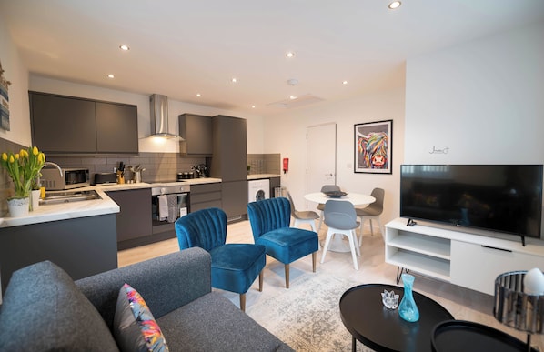 A modern living room with a grey sofa, blue velvet chairs, a flat-screen TV, and a dining area with a white table and chairs.
