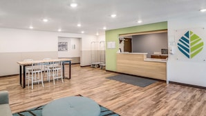 A modern reception area with a wooden desk, white chairs, a blue ottoman, and a green accent wall. - Woodspring Suites  West Des Moines (West Des Moines)