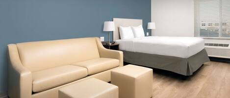 Quarto de hotel moderno com sofá bege, um banquinho combinando, cama com roupa branca e mesa de cabeceira com luminárias.