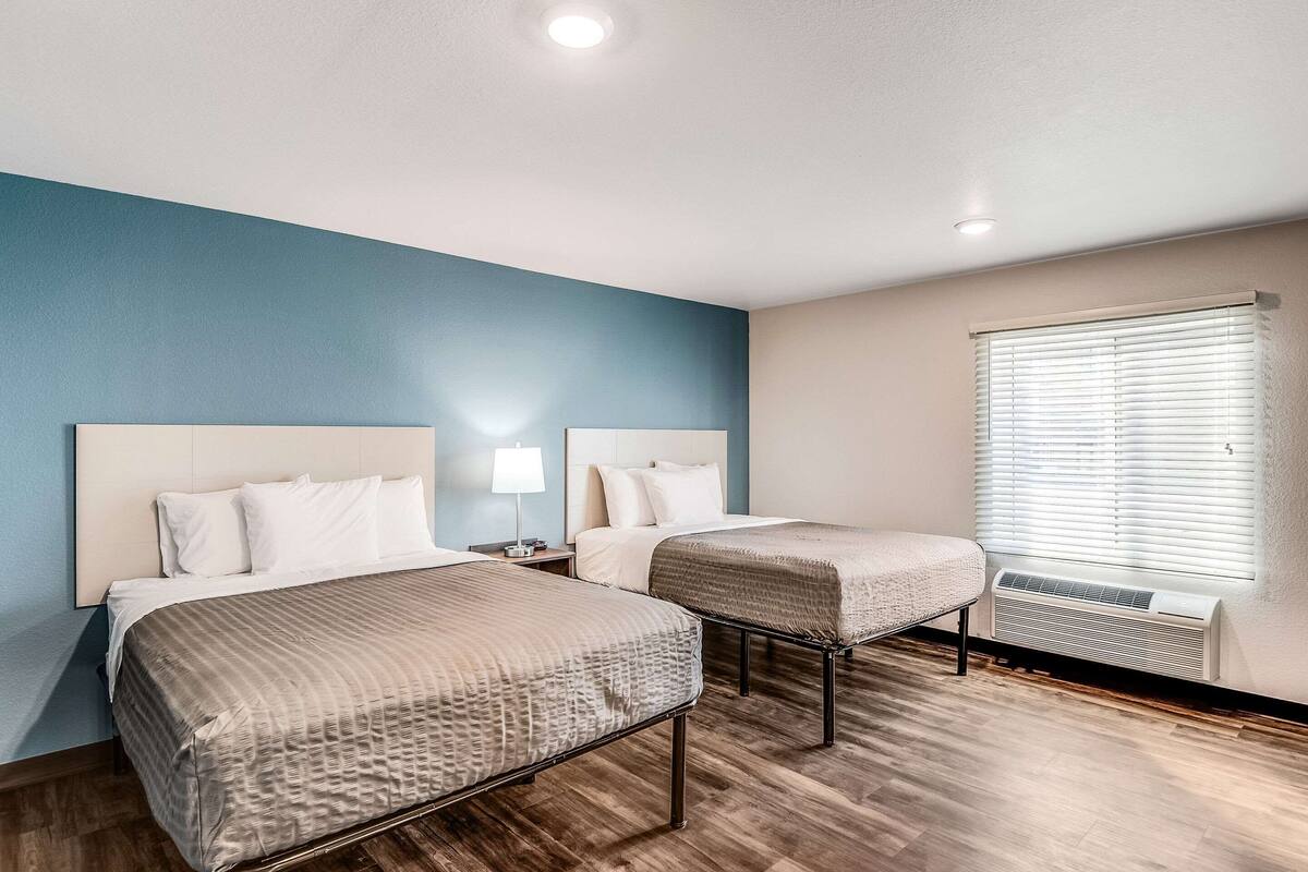 A hotel room with two beds, a nightstand, a lamp, and a window with blinds.