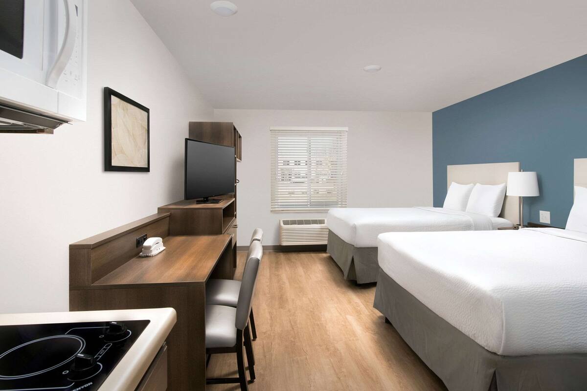 A hotel room with a kitchenette, two beds, a desk with a chair, a TV, and a window with blinds.