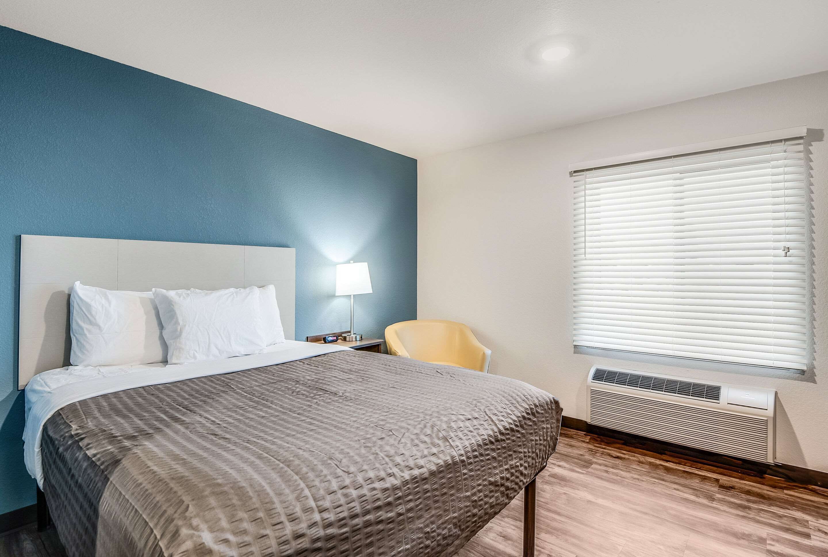 A hotel room with a bed, a chair, a lamp, and an air conditioning unit.