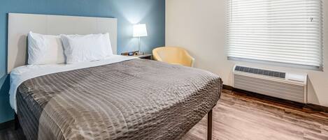 A hotel room with a bed, a chair, a lamp, and an air conditioning unit.