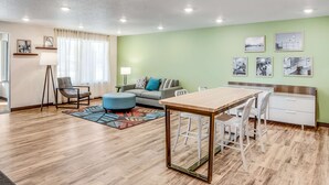 A modern living room with a wooden floor, a sofa, a blue ottoman, a dining table with chairs, and a kitchen area with a white cabinet. - Woodspring Suites  West Des Moines (West Des Moines)