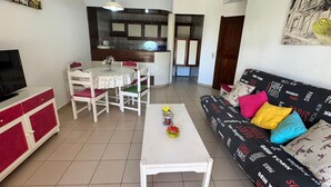 A living room with a TV, dining table, chairs, a sofa, and a kitchen area in the background. - Nautilus A (Quarteira)