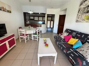 A living room with a TV, dining table, chairs, a sofa, and a kitchen area in the background.