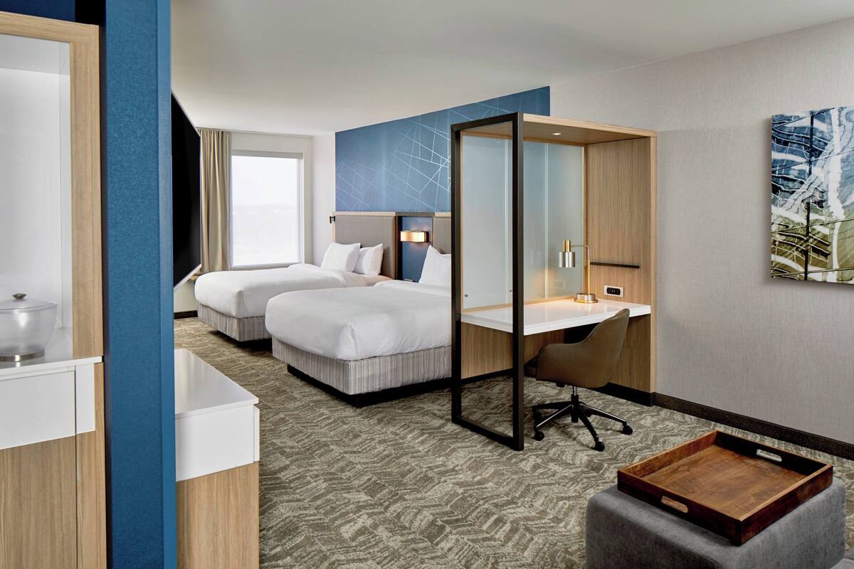 A hotel room with two beds, a desk, a chair, a small table, and a large window.