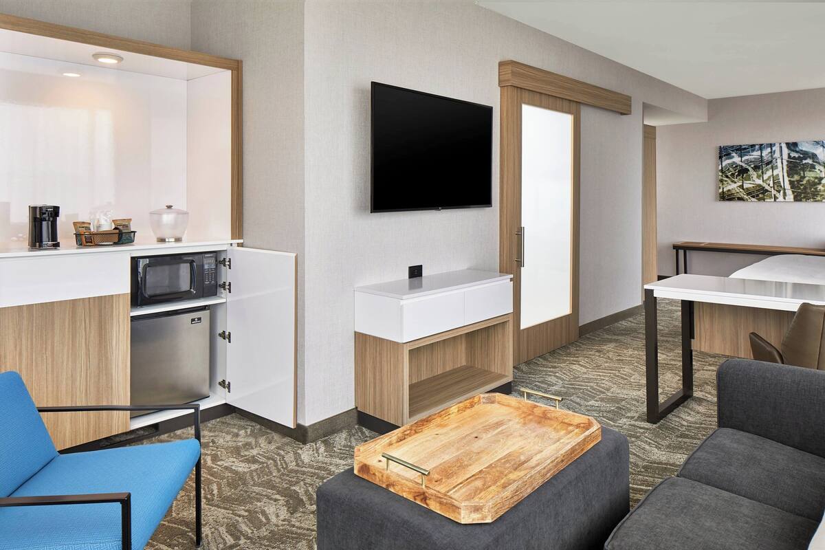 A compact hotel room with a kitchenette, a flat-screen TV, a dining area, and a seating area with a wooden coffee table.