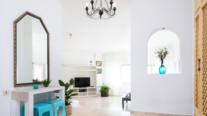 Un salon spacieux avec une console blanche, un grand miroir et un téléviseur. On y trouve également des plantes en pot et une fenêtre donnant sur un beau paysage.