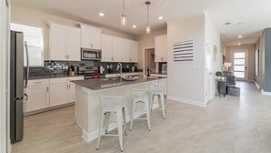 A modern kitchen with white cabinets, a central island with bar stools, and a dining area with a small table and chairs. - Ruby Falls Villa (Kissimmee)