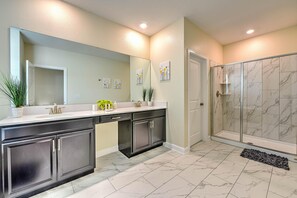 A modern bathroom with a large mirror, dark cabinets, a white countertop, and a shower with marble tiles. - Blooming Villa (Kissimmee)