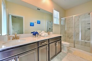 A modern bathroom with a large mirror, a white sink with gold fixtures, a glass-enclosed shower, and marble flooring. - Blooming Villa (Kissimmee)