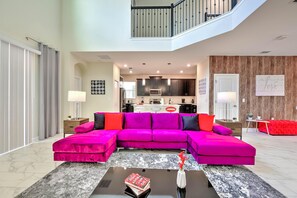 A modern living room with a vibrant pink sectional sofa, a glass coffee table, and a kitchen in the background. - Blooming Villa (Kissimmee)