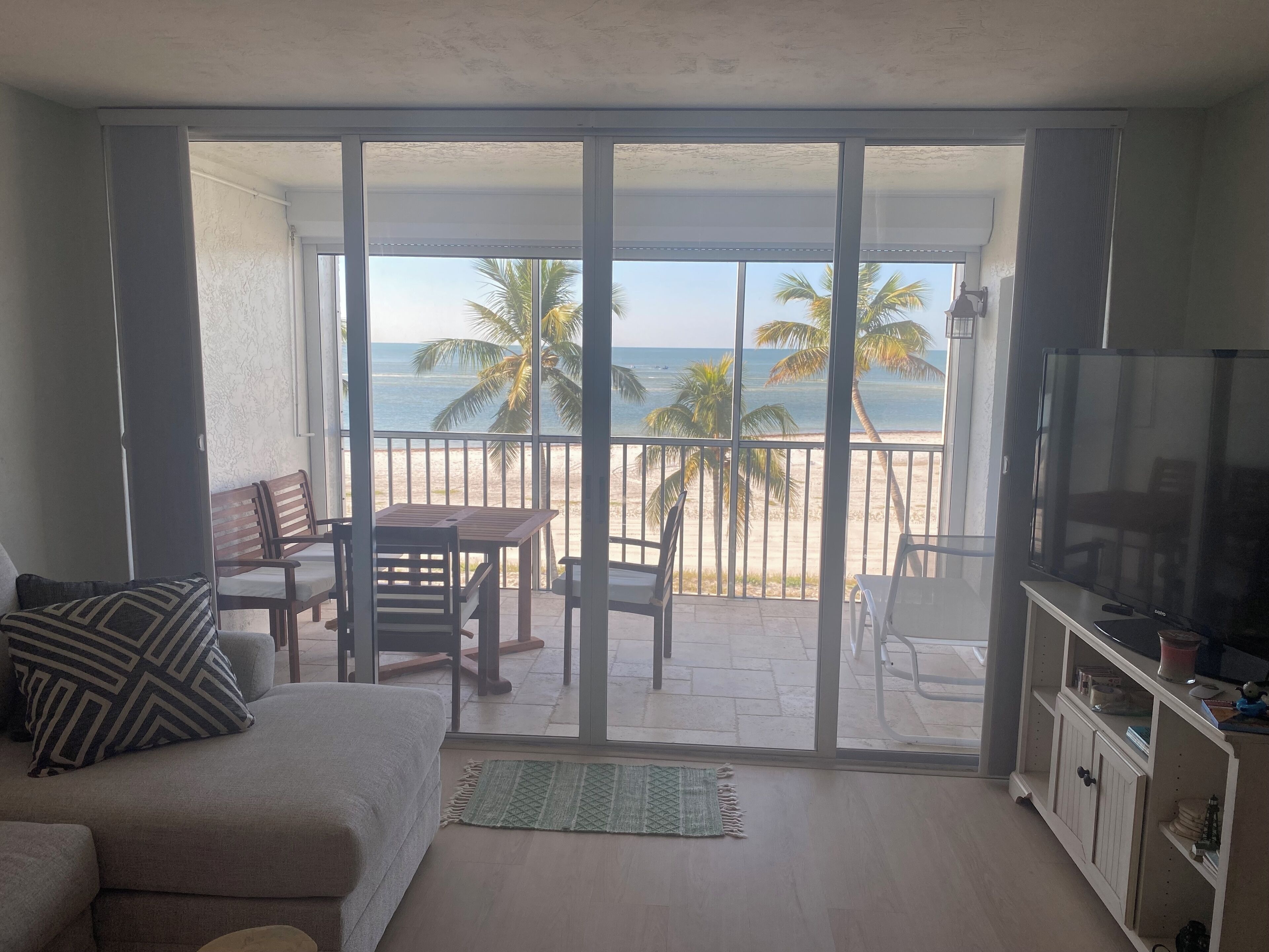 A living room with a sofa, a table, and chairs, a TV, and a sliding glass door leading to a balcony with a view of the beach and palm trees.