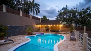 Ein Pool mit geschwungener Kante, umgeben von Steinplatten und einem weißen Zaun. Liegestühle und ein kleiner Barbereich mit Lichterketten sind vorhanden.