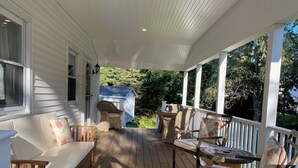 Une véranda couverte avec une terrasse en bois, des meubles en osier et un petit abri de jardin en arrière-plan.