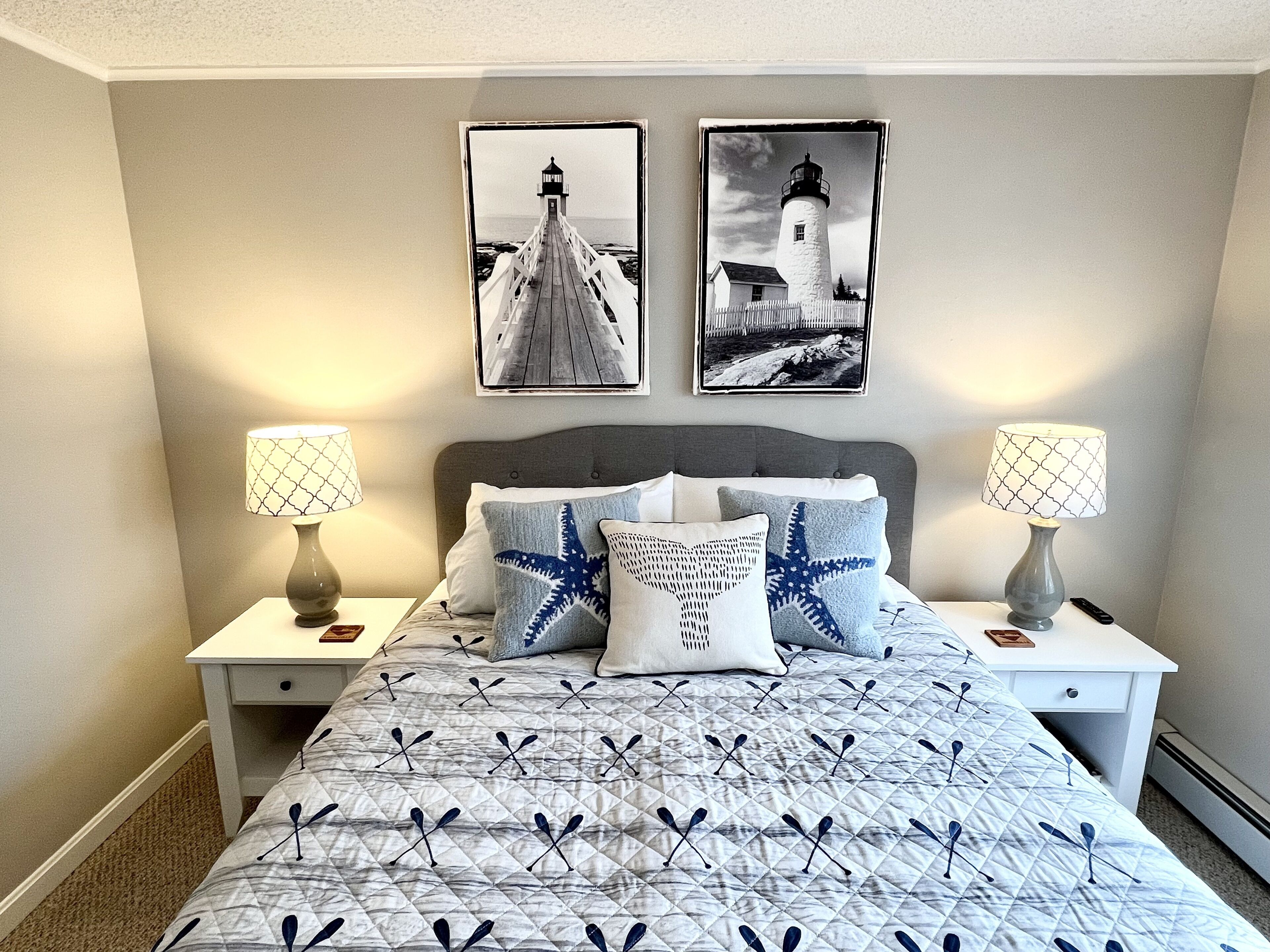 Une chambre à coucher comprenant un lit, deux tables de chevet, deux lampes fixées au mur, une tête de lit et deux tableaux représentant des phares.