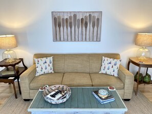 Uma sala de estar com um sofá bege, duas luminárias, uma mesa de centro com livros e uma tigela decorativa, e uma obra de arte na parede acima do sofá.