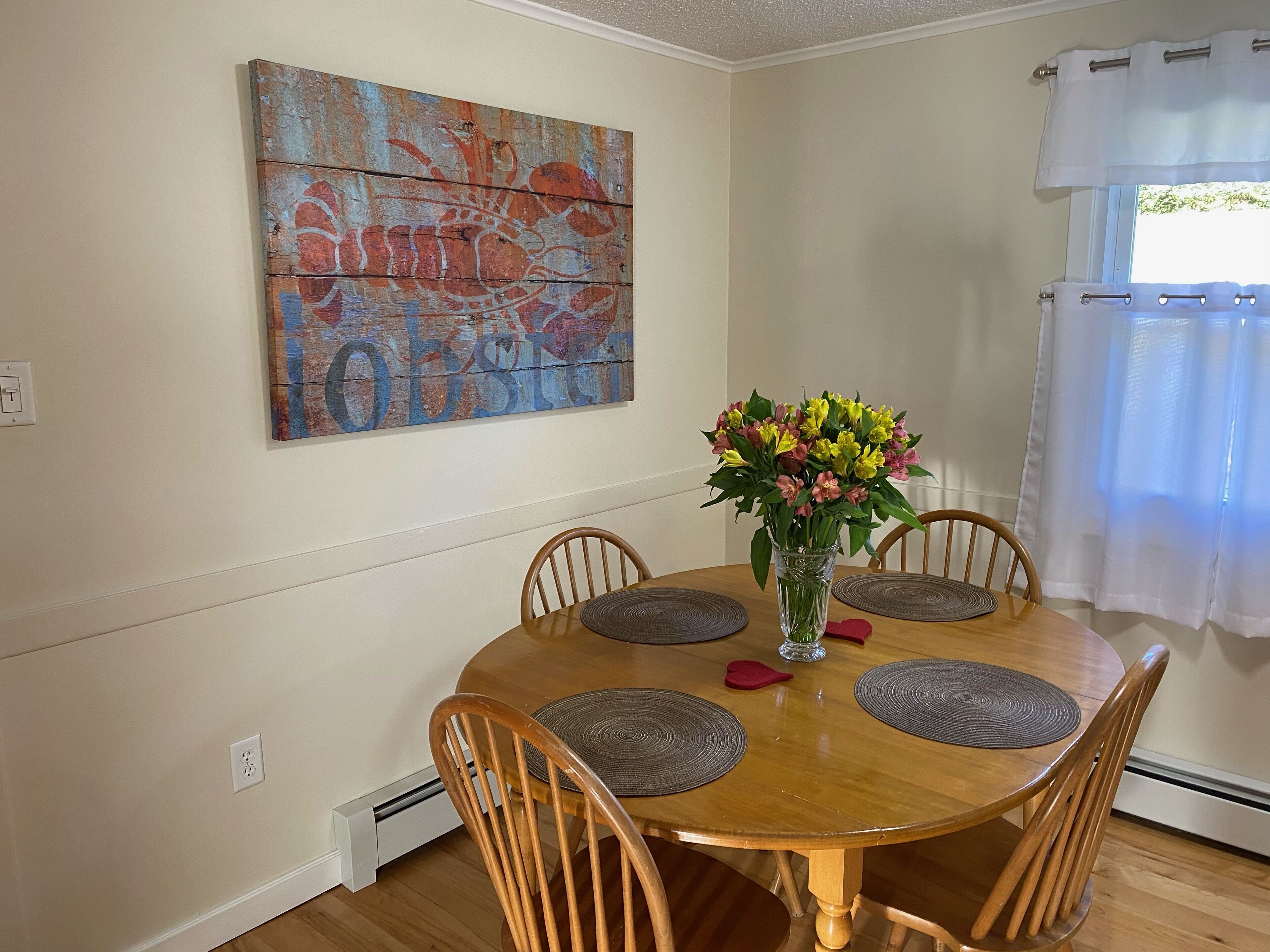 A dining room with a wooden table, four chairs, a vase with flowers, and a large abstract painting on the wall.
