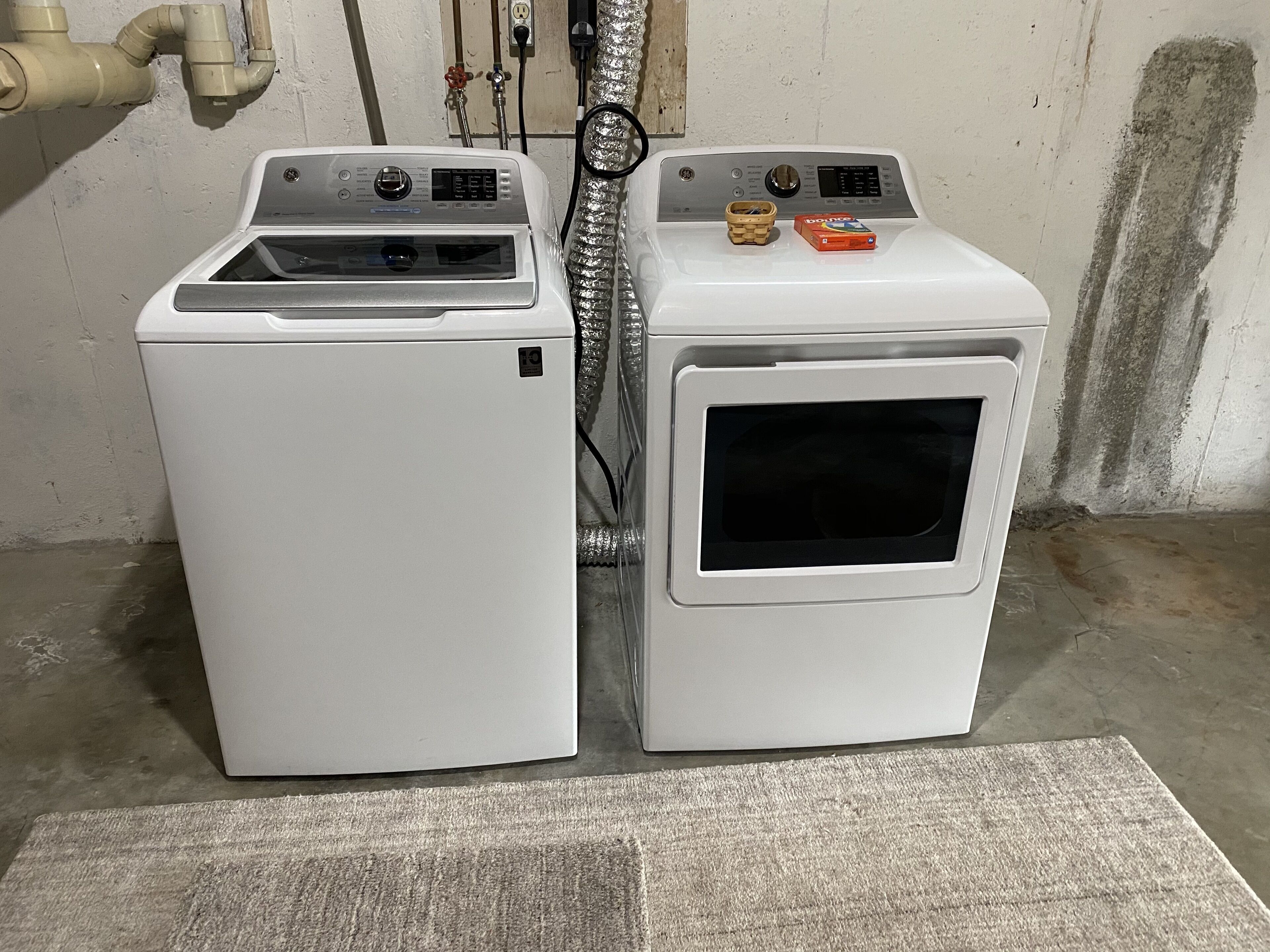 Deux appareils électroménagers blancs superposés, probablement une machine à laver et un sèche-linge, avec un câble d’alimentation visible et un petit récipient placé sur le dessus.