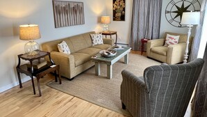 Un salon comprenant un canapé, deux fauteuils, une table basse et une lampe. Un tableau est accroché au mur, et il y a une décoration en forme de boussole.