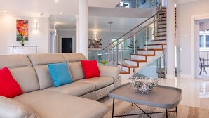 A modern living room with a beige sectional sofa, a glass coffee table, and a staircase with wooden steps and glass railings.