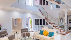 Un vaste espace de vie agrémenté d’un escalier moderne, d’un canapé blanc orné de coussins colorés et d’une table basse en verre.