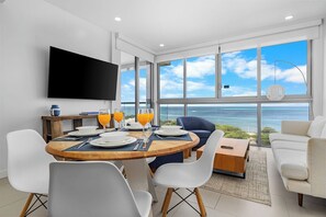 A modern living room with a large window, a flat-screen TV, and a dining area with breakfast set.