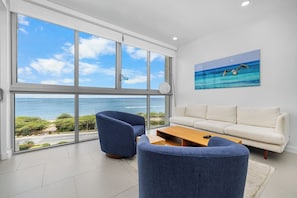 A modern living room with a large window overlooking the sea, a white sofa, blue armchairs, and a wooden coffee table. - Real Retreat with Ocean View. 2BDR condo for 5 (Eagle Beach)