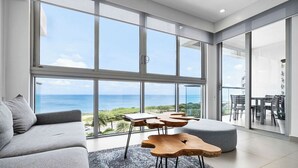 A modern living room with a large window overlooking the ocean, a gray sofa, and unique wooden tables. - Peace and Light, Ocean View Condo on the 5th floor (Eagle Beach)