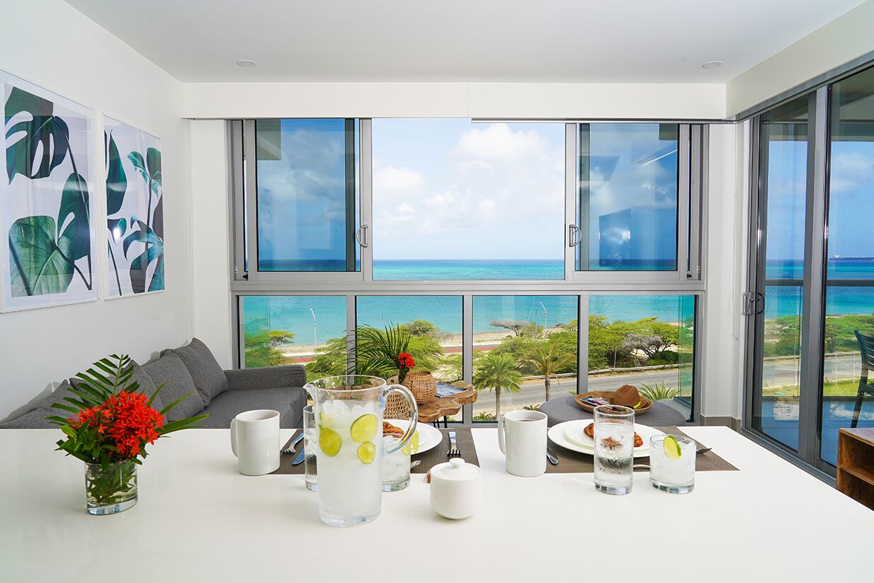 A modern dining area with a table set for breakfast, large windows with a view of the ocean, and tropical artwork on the wall.