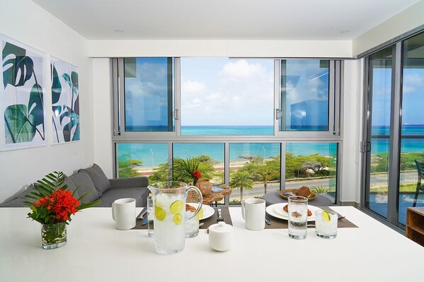 A modern dining area with a table set for breakfast, large windows with a view of the ocean, and tropical artwork on the wall.